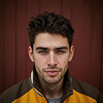 Young man with dark curly hair and a light beard wearing a yellow and black jacket against a brown wooden background.